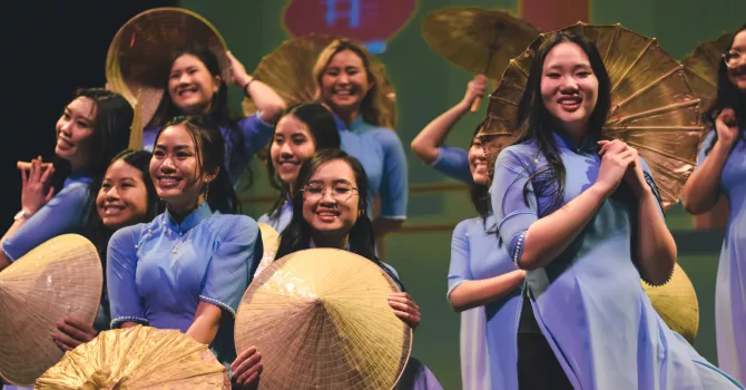 Vietnamese students on stage wearing purple & smiling, some with straw hats