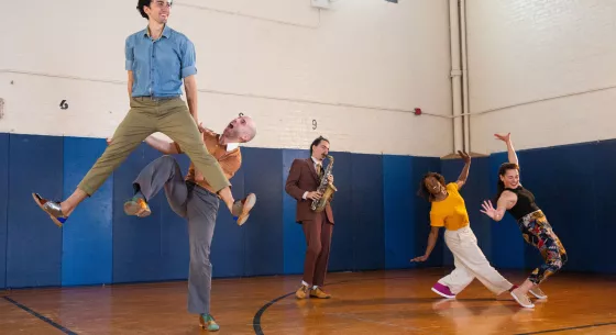 A pair of Swing dancers and a saxophonist playing in the backgound. One couple is lifting their partner in the air