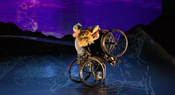 Shadowy images of Rodin sculptures hang in a lilac-streaked sky. Laurel Lawson lifts Alice Sheppard onto her lap. Alice looks down at the shadowy lines on the deck of the ramp; her wheels hang and her crown of golden curly hair shines in the light. Laurel holds Alice and turns her chair. 