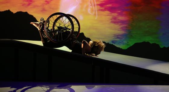 As the sky transitions from gold to amber to red, green and blue, Alice Sheppard, a light-skinned Black woman, slides in a spider position on her stomach down the shiny ramp. The shadow of her wheelchair is visible beneath; her curly hair glows. Photo credit: MANCC / Chris Cameron.
