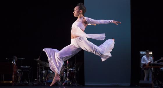 A female dancer wearing white leaps forward on stage.