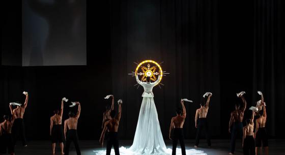 A group of dancers wearing black with their back to the audience face a dancer twice as tall as them wearing a long white dress holding a glowing yellow circle with a star design in the middle. 