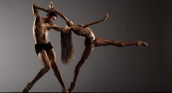 Two dancers wearing brown and black appear in front of a gray backdrop. One dancer extends a leg straight to the right while bending backward toward the second dancer, who stands at a slight angle with one leg bent at the knee, holding onto the arm of the other dancer.
