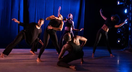 A group of 5 dancers wearing black clothing appear onstage with cobalt blue lighting. 3 dancers stand facing the camera with their arms extended upward and parallel to the floor while 1 dancer kneels, facing the curtain.
