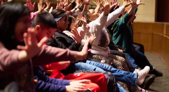 Kids sit in the audience and enthusiastically wave their hands in the air.