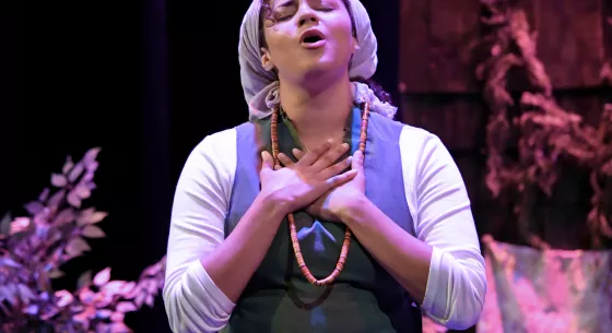 Woman in purple dress and headscarf holds her hands over her heart and lifts her head, eyes closed