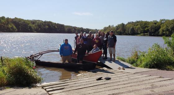 A group of 14 students stand on a dock behind a large canoe floating in a lake.