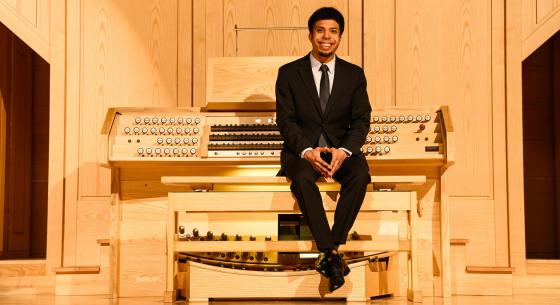 An organist with dark hair wearing a black suit and tie sits in a recital hall with their back towards a wooden organ console and fingers intertwined and placed on their knees.