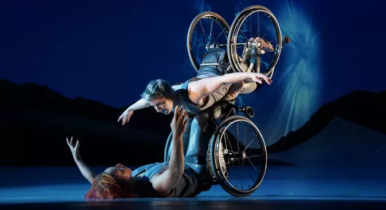 Laurel Lawson, a white woman, is flying in the air with arms spread wide, wheels spinning, and supported by Alice Sheppard. Alice, a light-skinned Black woman, is lifting from the ground below. They are making eye contact and smiling. 