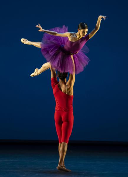 A male dancer wearing a red costume holds a female dancer wearing a purple costume above his head as she poses.