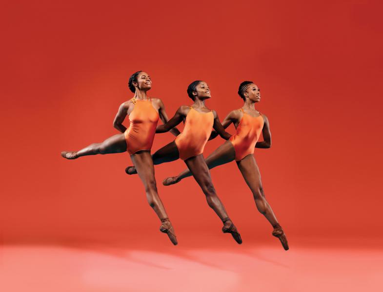 Three female dancers perform together in front of an orange background.