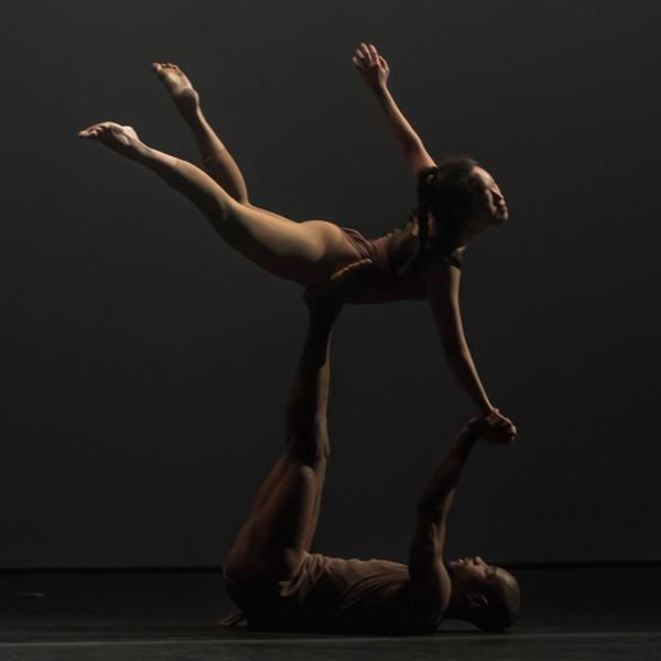 Male dancer is on the floor, female dancer is lifted into the air with his foot on her stomach, her legs back and her hand lowered to reach his extended hand 