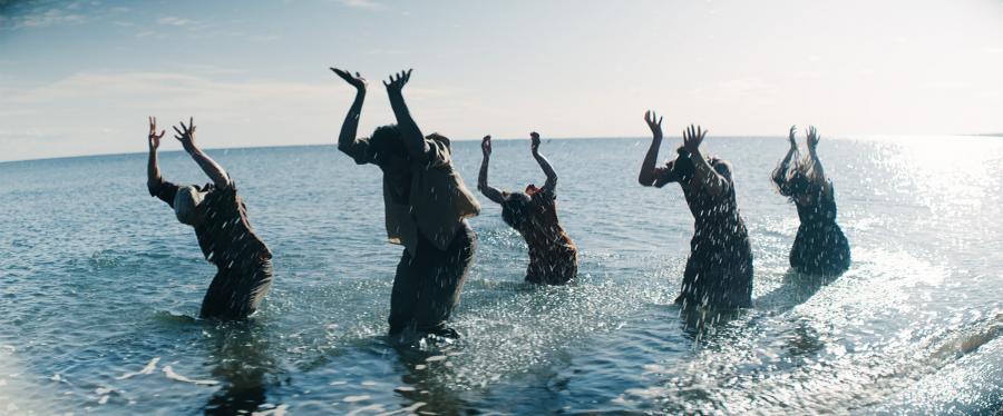 Dancers perform in a body of water with their hands in the air.