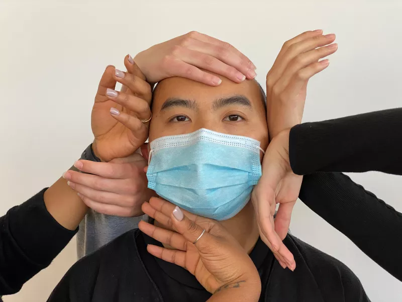 Film still - closeup of a person in a face mask surrounded by hands 