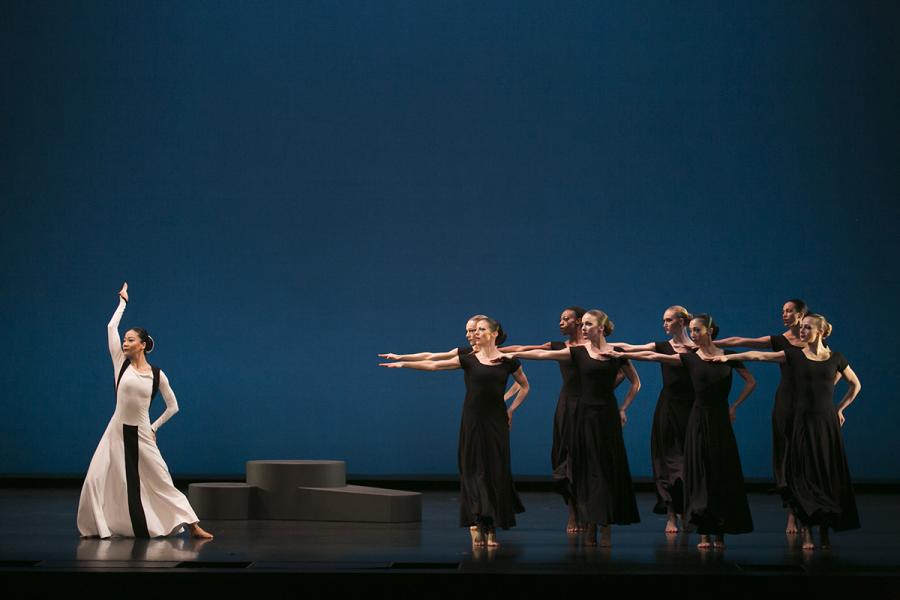 Several dancers dressed in black point their arms towards one dancer all in white
