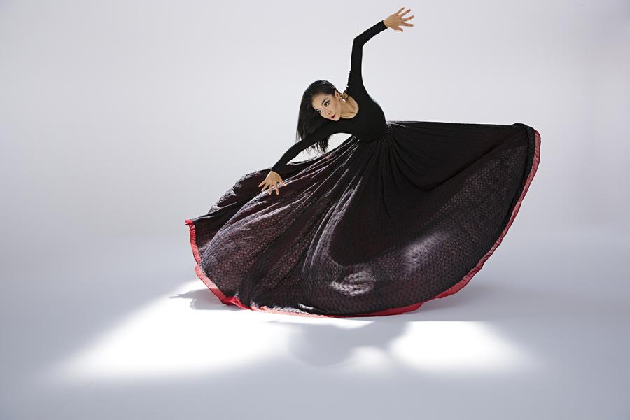 A woman dances with the skirt of her dark dress flared wide and her arms in the air.