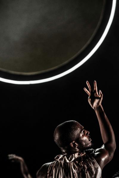 A larger sphere who's cirfumference is outlined with a white light, hangs from the top left corner of the retangular photo. The white glow refelcts off of a muscular, short haired dancer in the bottom right corner, who is posing with their arms held above their head. 