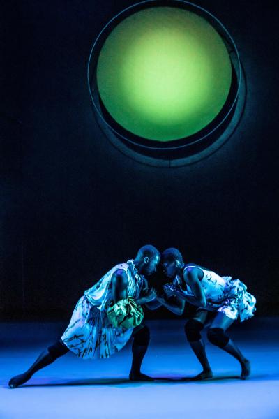 Two dancers, touching hands and leaning in towards eachother in a lunge position on a blue lit stage floor. Above hangs a neon green, glowing sphere of light.