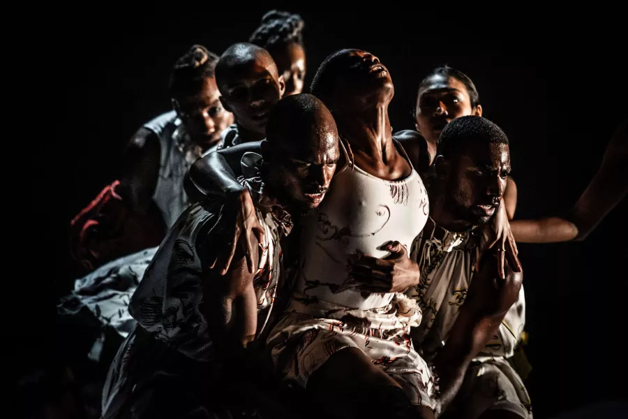 Being lifted from beneath each armpit and support from behind, a dancer in distress is being carried across the stage by a group. 