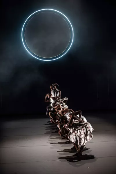 A large sphere who's cirfumference is outlined with a blue light, glows upon a vertical lineup of dancers, each striking poses of various heights with the lowest being in the front, and the tallest (standing) in the back. 