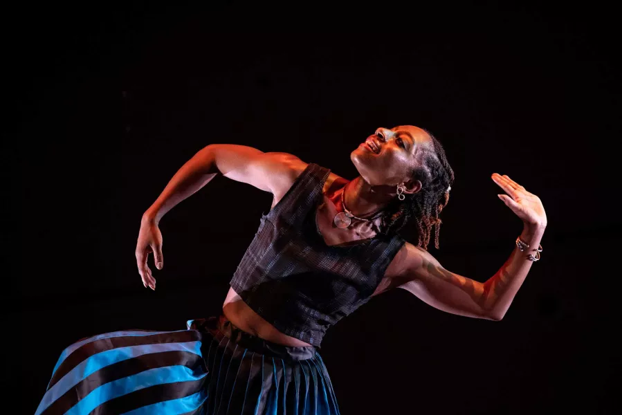 A dancer kneels on stage, with their head turned to the right, and each arm positioned in a half bent pose.