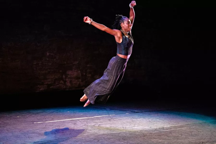 A dancer strikes a jumping position in the air, with both legs held tightly underneath their torso, and both arms extended over their head.