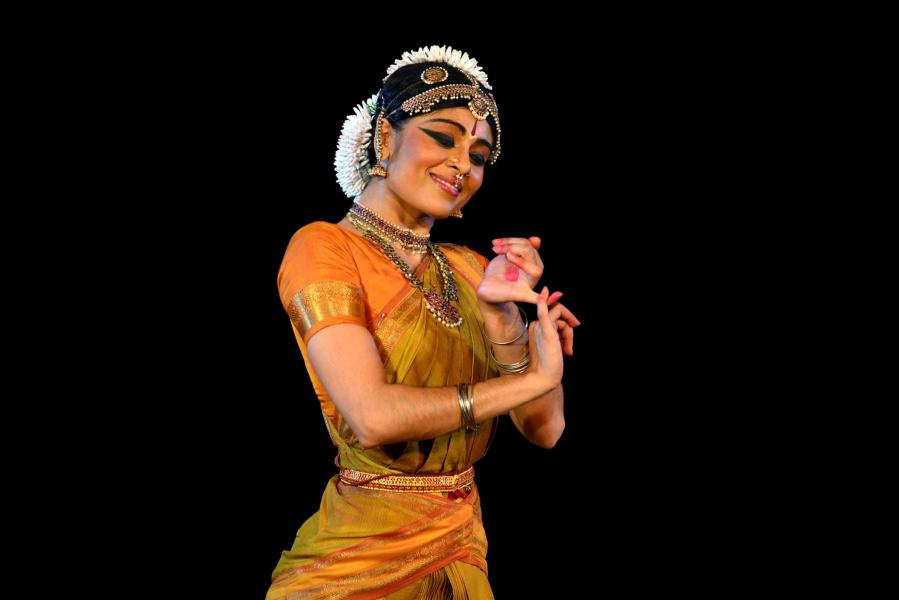 A dancer wearing an orange and gold sari with traditional jewels and accessories, forms a gesture with their hands while having a smiling expression on their face.