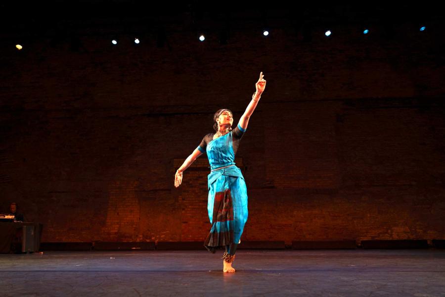 A dancer wearing a blue sari, is standing center stage, striking a pose with their left arm extended above their head.
