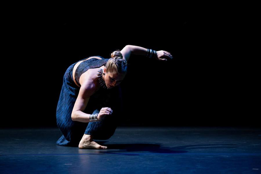 A dancer strikes a pose in a lunging position, with their head low to the ground, and arms extended to either side of their body, while wearing a dark blue shirt and pants that matches the dark lighting of the stage and backdrop.