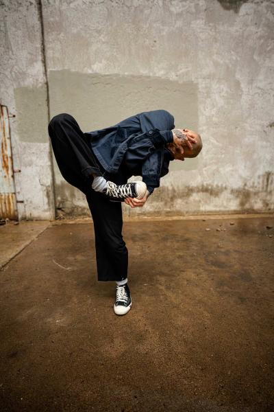 A dancer stands in a concrete alleyway, wearing a denim shirt and pants, paired with black converse. The dancer stands on one leg, while the other leg is being held in the air by his opposite arm. The remaining arm is being used to cover their face with their hand.