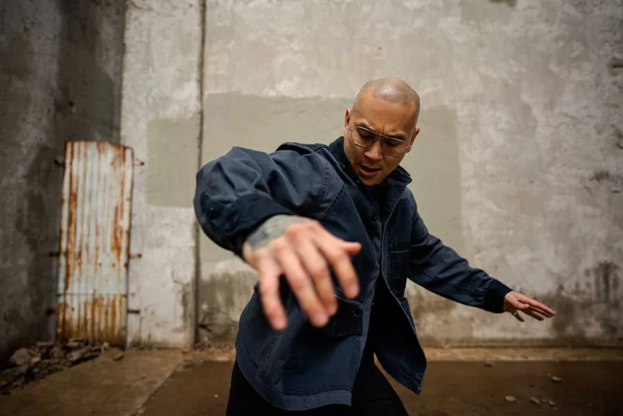 A dancer stands in a concrete alleyway wearing a blue jacket, with both arms extended out to either side of their body.