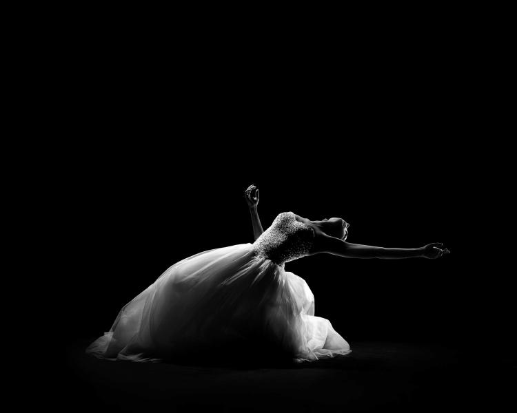 A dancer wearing a long white dress bends their knees and arches their back so their upper body is facing the sky with their arms are out to the side.