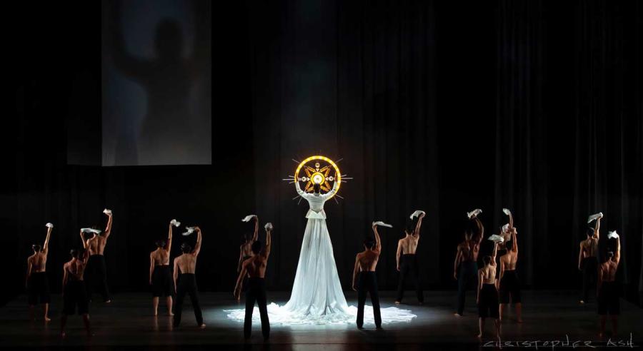 A group of dancers wearing black with their back to the audience face a dancer twice as tall as them wearing a long white dress holding a glowing yellow circle with a star design in the middle.