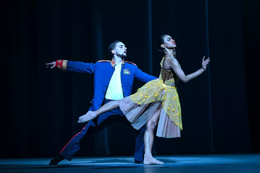 A dancer in a yellow dress strikes an arabesque pose, while a dancer behind holds them by the waist. 