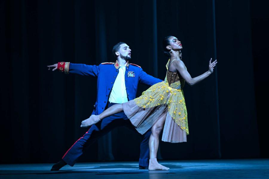 A dancer in a yellow dress strikes an arabesque pose, while a dancer behind holds them by the waist.