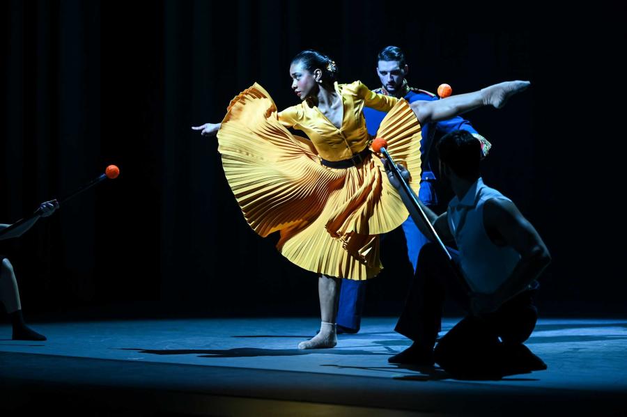 A dancer in a yellow dress strikes a one-legged pose with her remaining arms and legs extended. Another dancer is running towards her from behind.