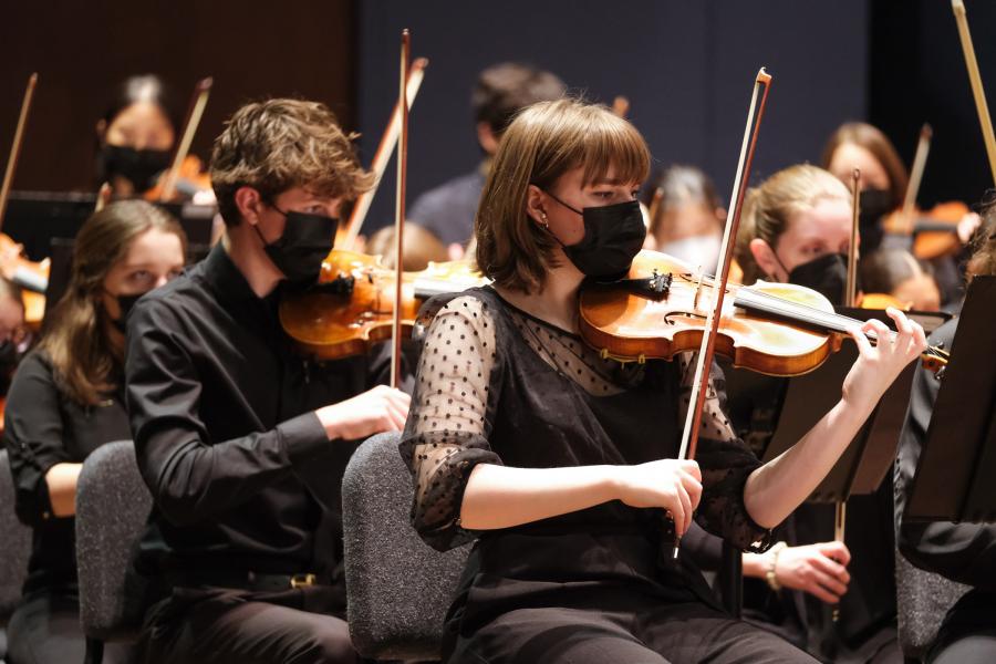 A group of musicians wearing black outfits and black masks covering their mouths hold violins on their left shoulder and hold the bow in their right hand.