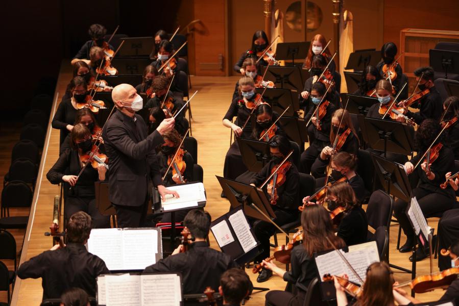 One person stands in front of a group of musicians who are seated facing them. All of the musicians are in rows playing wood violins. The person standing is holding a baton and appears to be directing the group.