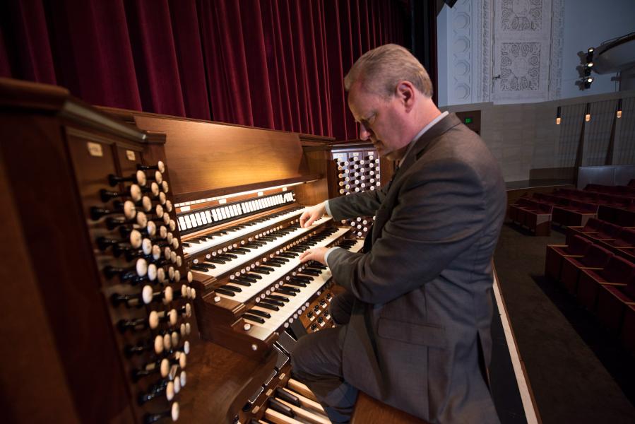 An organist wearing a dark grey suit faces a large, wooden organ and looks at the keys. Their left hand is on the second row of keys and their right is on the third.
