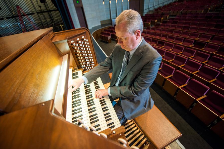 A birds-eye view image reveals a person with grey hair sitting at an organ with their hands on the second and third rows of keys. There are rows of maroon seats in the background.