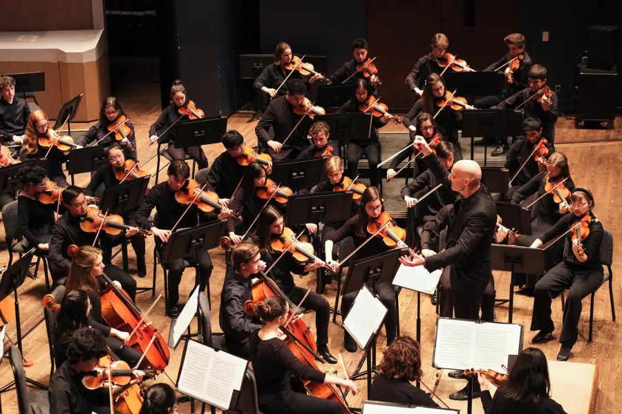 Rows of young people playing various woodwind instruments are seated around a person standing who appears to be directing the group.