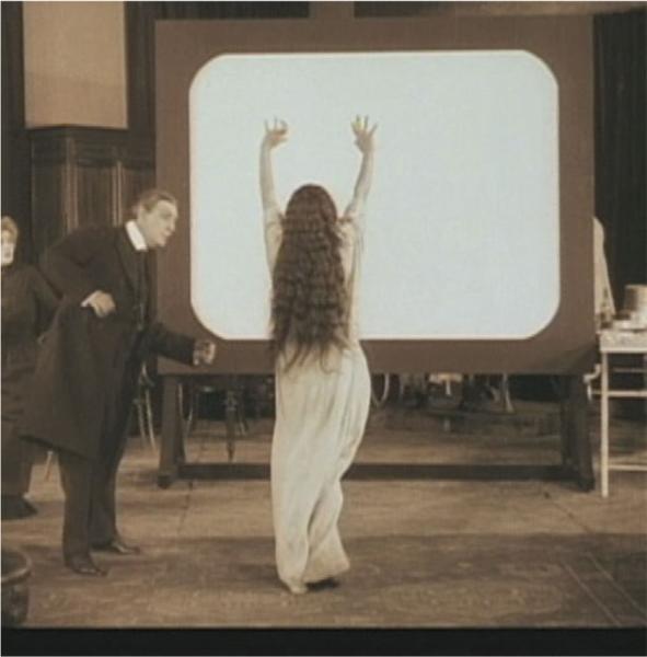 Sepia toned image of the back of woman in whit gown and long hair with arms raised faces a white screen. A man in a suit stand close to her.
