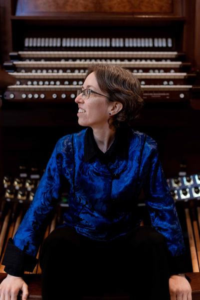 An organist wearing a deep blue button down with short, dark brown hair and glasses sits on a bench in front of a large, wooden organ console with their head turned to the right.