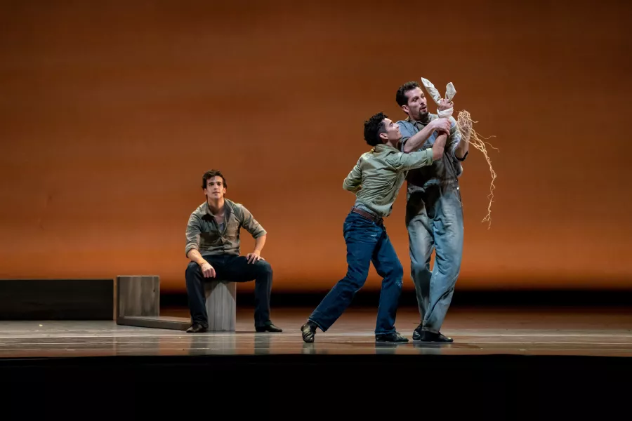 Three dancers are on an orange lit stage. One sits on a wooden stool upstage, while the the other two stand downstage, clutching each others wrists.