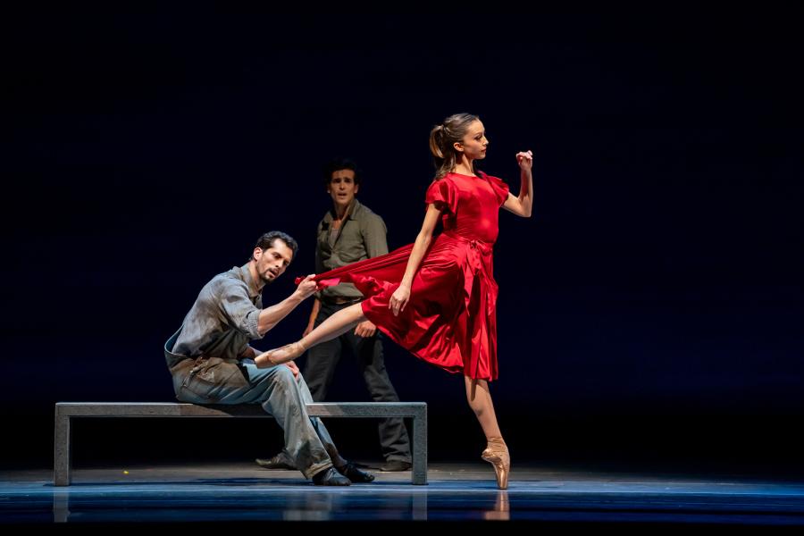 Two dancers, one sitting on a long, narrow bench, while the other stands, both admire a dancer forefront of them who is standing in an arabesque pose en pointe.