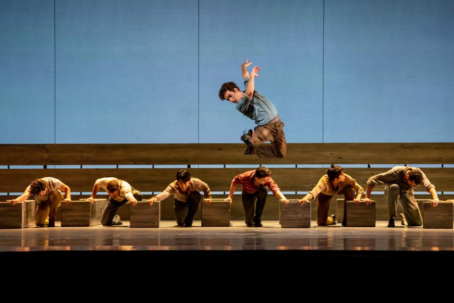 A dancer center stage strikes a jumping pose in the air, while upstage six dancers kneel, with a wooden box on either side of them that they each are holding onto.