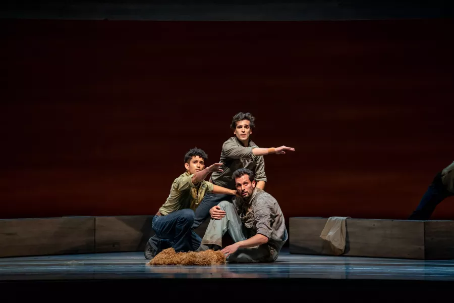 Three dancers are center stage. One sits, one kneels, and one squats. Those kneeling and squatting have their arm pointing out in front of them. The sitting dancer looks in the direction being pointed too.