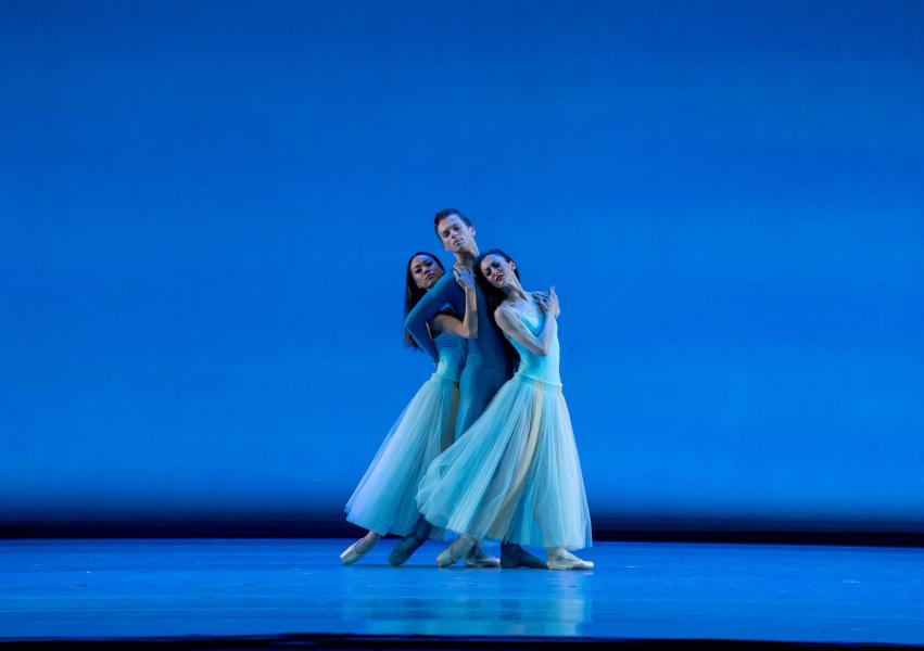 Three dancers stand center on a blue lit stage while the backdrop, matches the blue costumes they all wear. The dancers stand close to each other in a horizontal line, each embracing the one next to them.