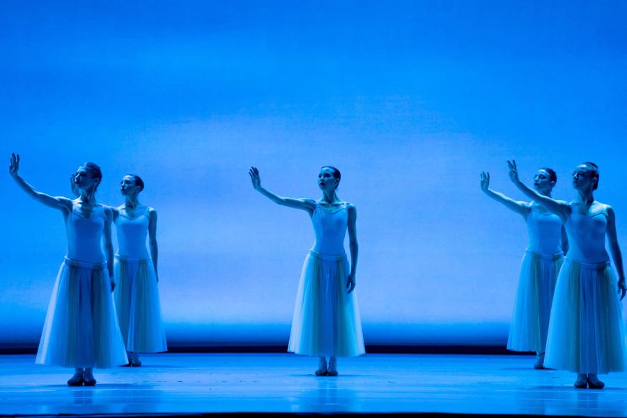 Three vertical rows of dancers are lined up on a blue lit stage and matching backdrop. Each dancer strikes the same pose: standing, with their right arm extended to a shoulder-height.