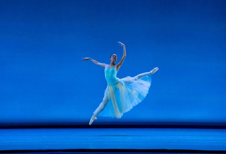 A dancer strikes a leaping position, centerstage in front of a blue lit backdrop. The dancers hind leg is extended to a waist height position, while the other is underneath their torso, Both of the dancers arms are above their head.